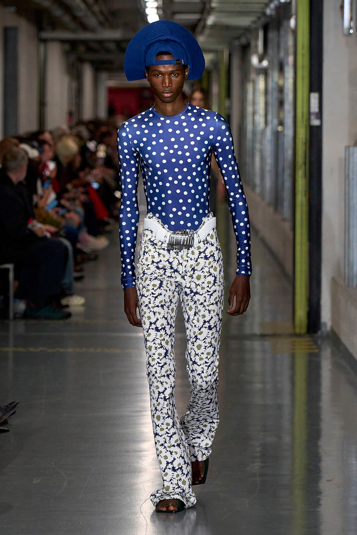 Male model walking a runway in a blue polka-dot bodysuit, white floral bell-bottom pants, a silver belt, and a large cobalt hat.