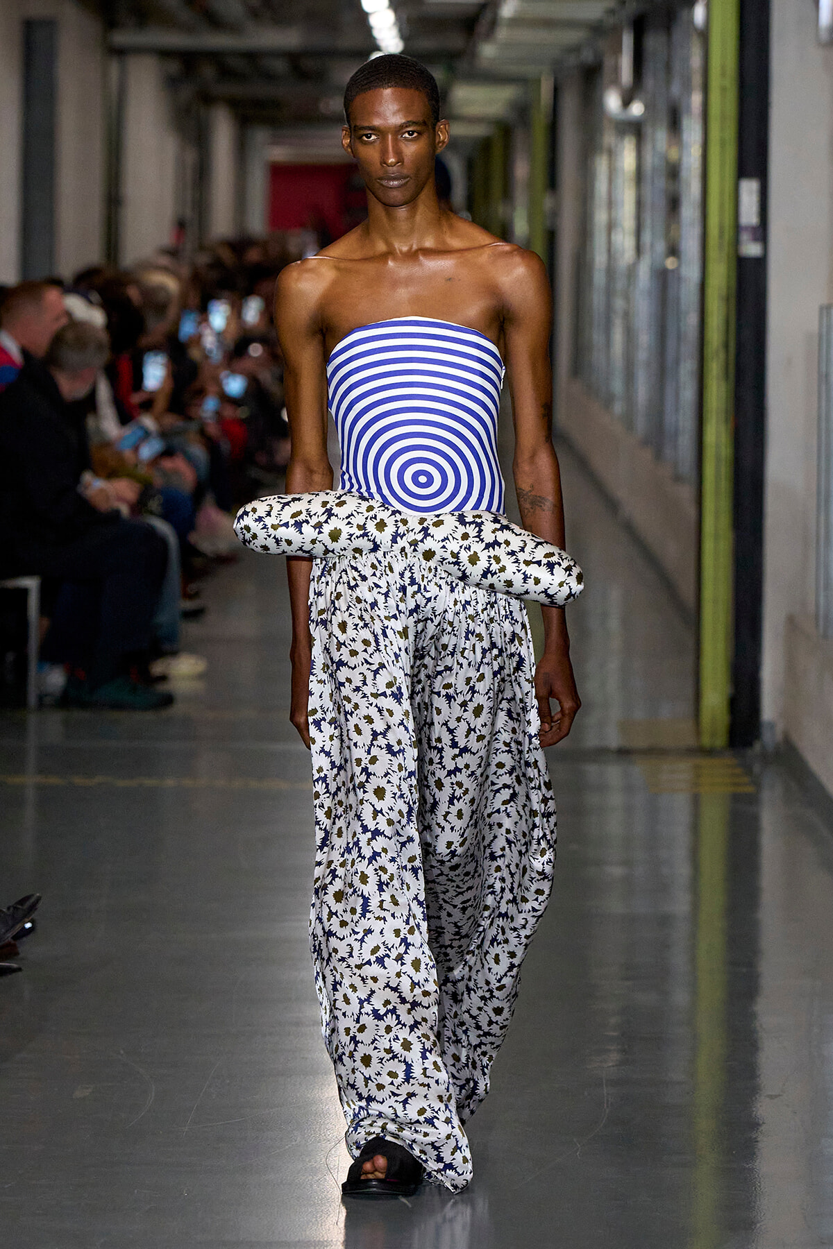 Runway model wearing a blue-and-white striped strapless top with oversized floral-print pants and a large daisy-pattern wrap at the waist