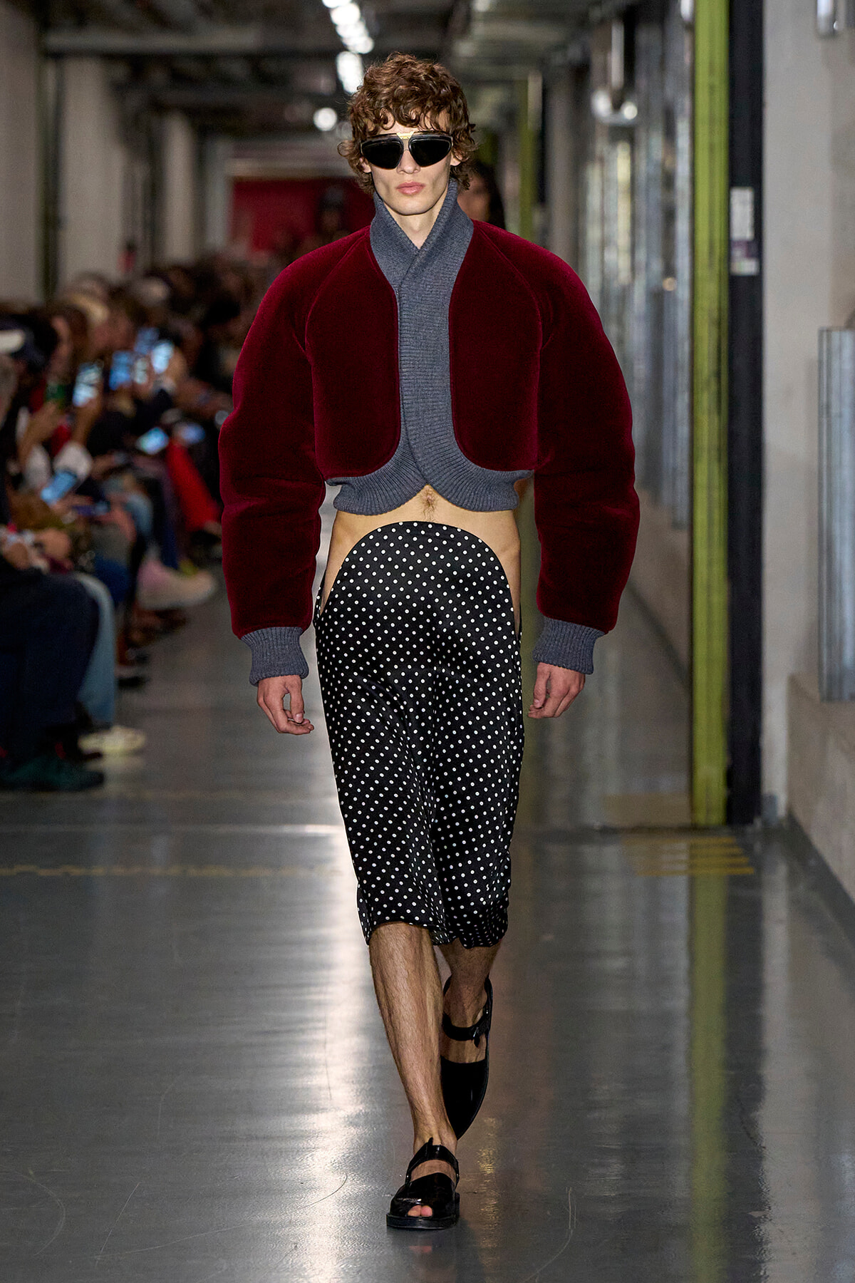 Male model on a runway wearing a cropped burgundy fleece jacket over a gray knit top, black-and-white polka-dot pants, sandals, and sunglasses.