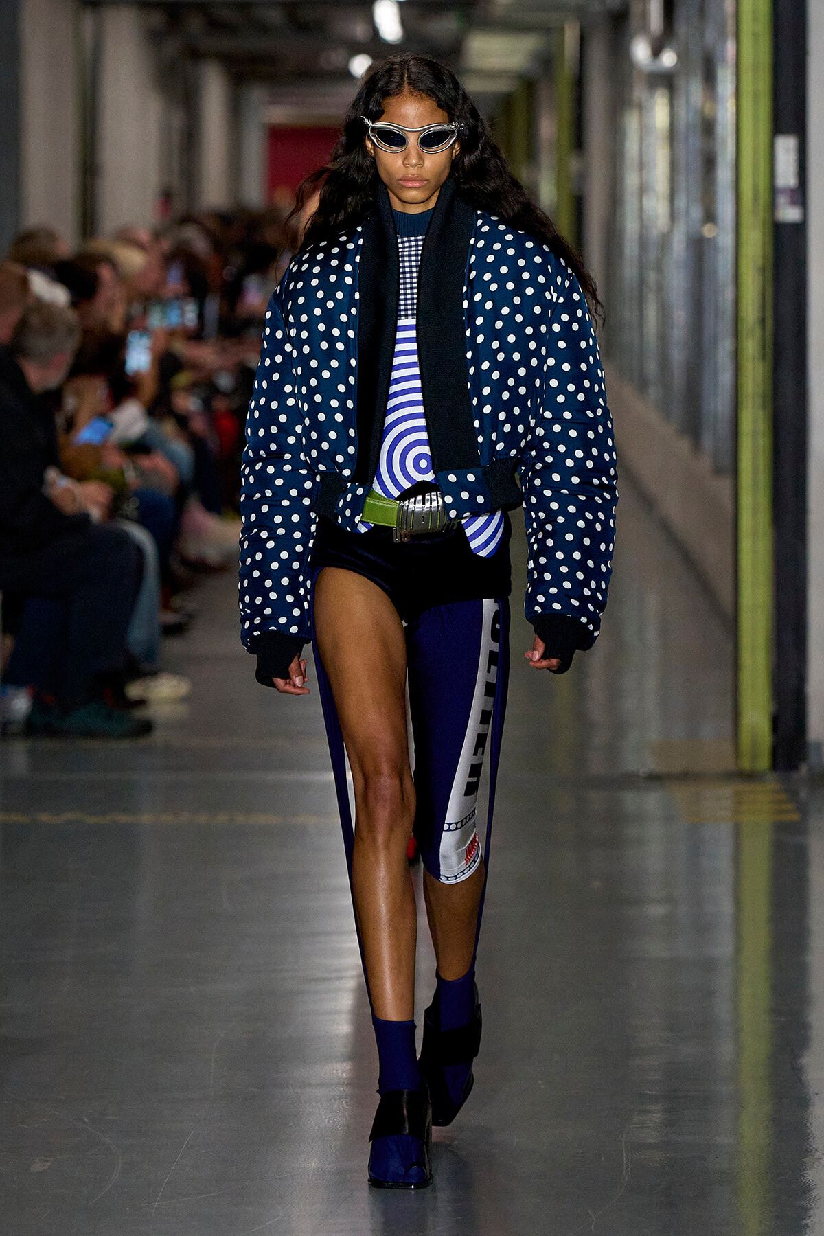 Model walking a runway in a blue polka-dot puffer jacket, white sunglasses, and athletic-inspired navy outfit with bold belt and patterned leg panel.