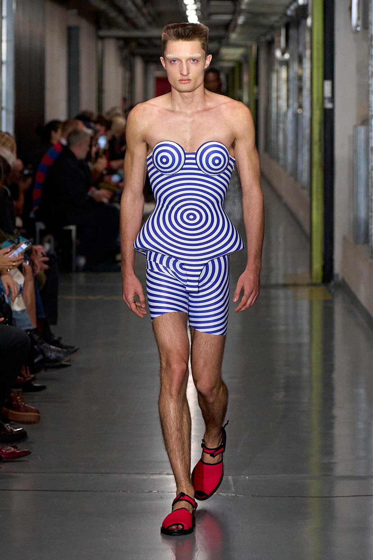 Male model walking a fashion runway in a blue-and-white spiral striped strapless bodysuit with circular chest cups and peplum, red sandals visible behind.