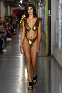 Model walking the runway in a gold sequin bikini with cutout sides and black sandals, backstage-style corridor in the background.