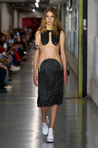 Model walks a runway in a black polka-dot cut-out top and matching cut-out midi skirt, paired with white ankle boots; audience blurred in the background.
