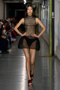 Runway model wearing a sheer black mesh dress with a high neck, paired with burgundy sandals, walking the catwalk.