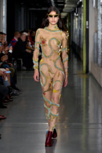 Model walks the runway in a sheer nude bodysuit with turquoise snake-like embroidery and floral appliqués, paired with burgundy ankle boots and yellow sunglasses.