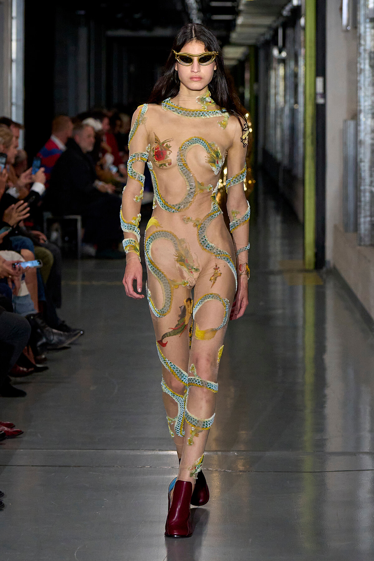 Model walks the runway in a sheer nude bodysuit with turquoise snake-like embroidery and floral appliqués, paired with burgundy ankle boots and yellow sunglasses.