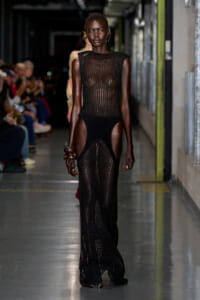 Model walks the runway in a sheer black mesh gown with high-cut sides; audience blurred in the background.