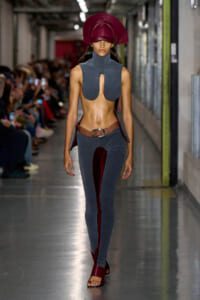 Model walking a runway in a gray sleeveless high-neck top with a front keyhole cutout, matching gray pants with red inner lining, brown belt, and a burgundy headpiece (avant-garde fashion)