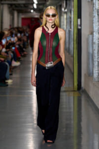 Model walking runway in a color-block burgundy and green corset top, wide black pants, silver belt, and futuristic sunglasses.