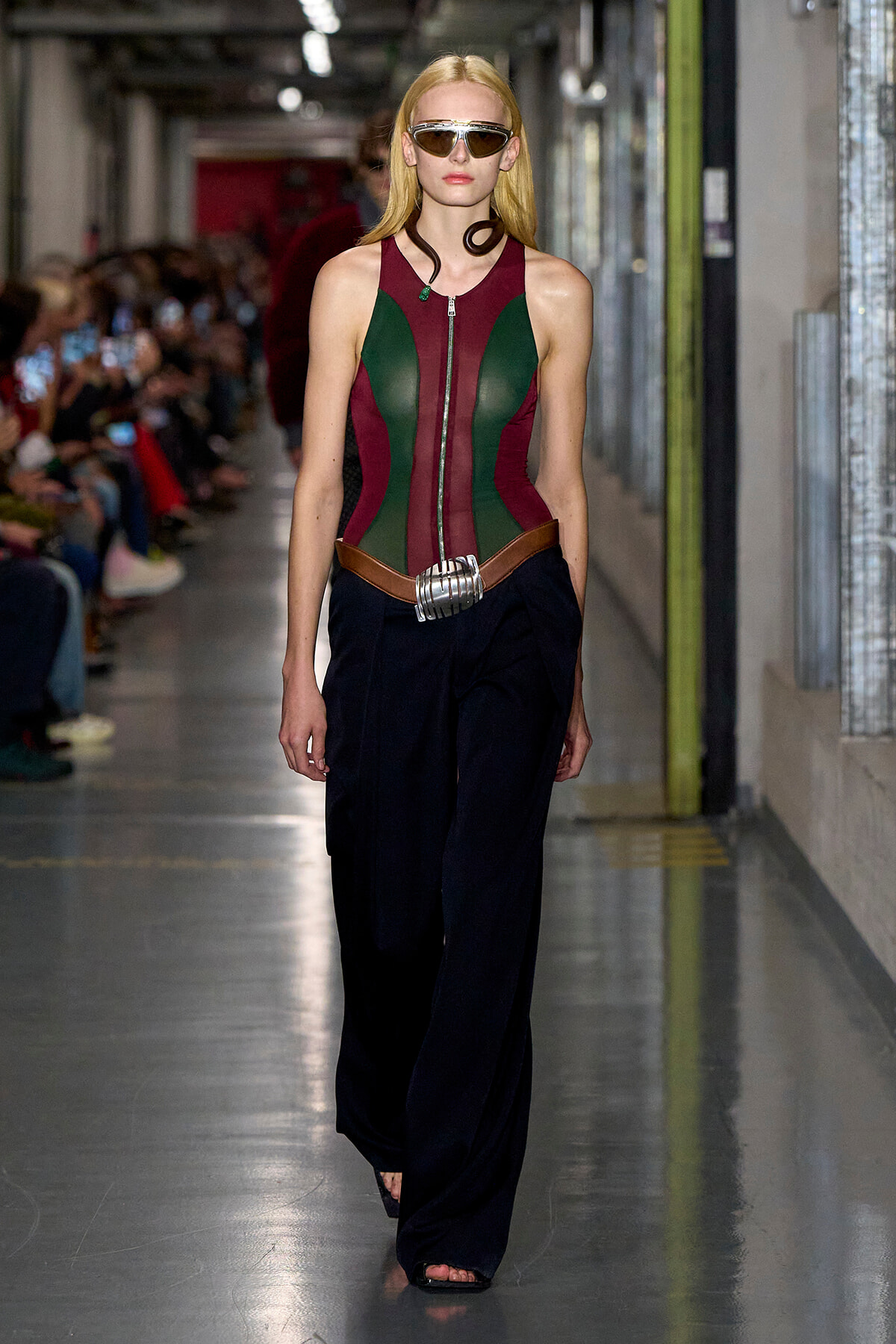 Model walking runway in a color-block burgundy and green corset top, wide black pants, silver belt, and futuristic sunglasses.