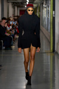 Model on a fashion runway wearing a black sculpted blazer dress with a high neck, red beret, polka-dot headband, and gold sunglasses with black ankle boots.