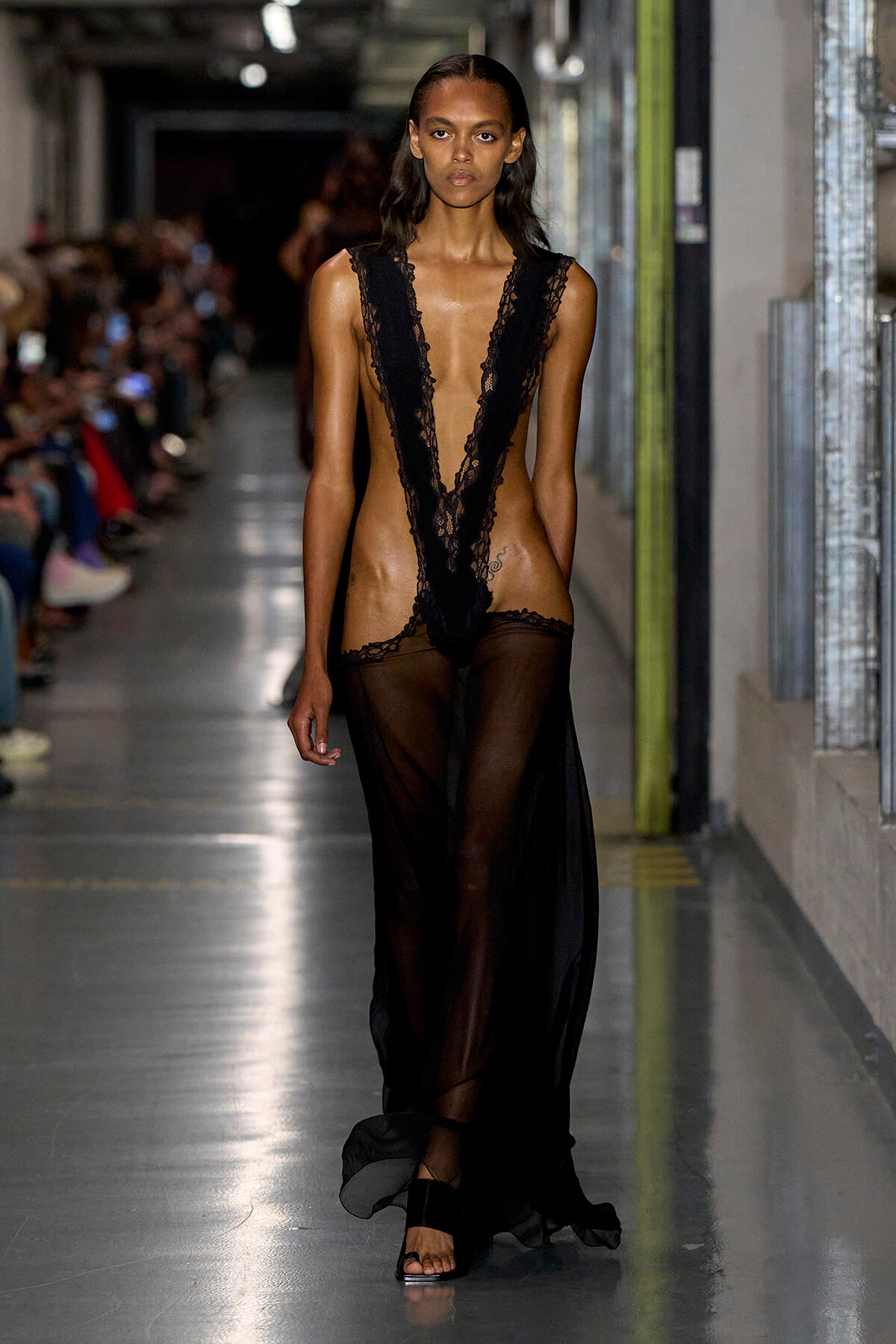 Model walks the runway in a black lace plunging V-neckline dress with a sheer skirt.
