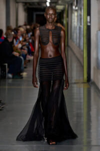 Model walks the runway in a black sheer two‑piece gown with a striped cutout bodice and flowing skirt.