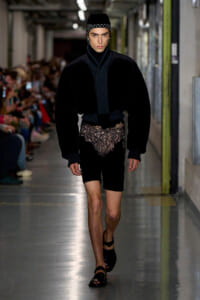 Male model walking a runway in a black velvet ensemble with lace shorts and a studded cap; audience blurred in the background.