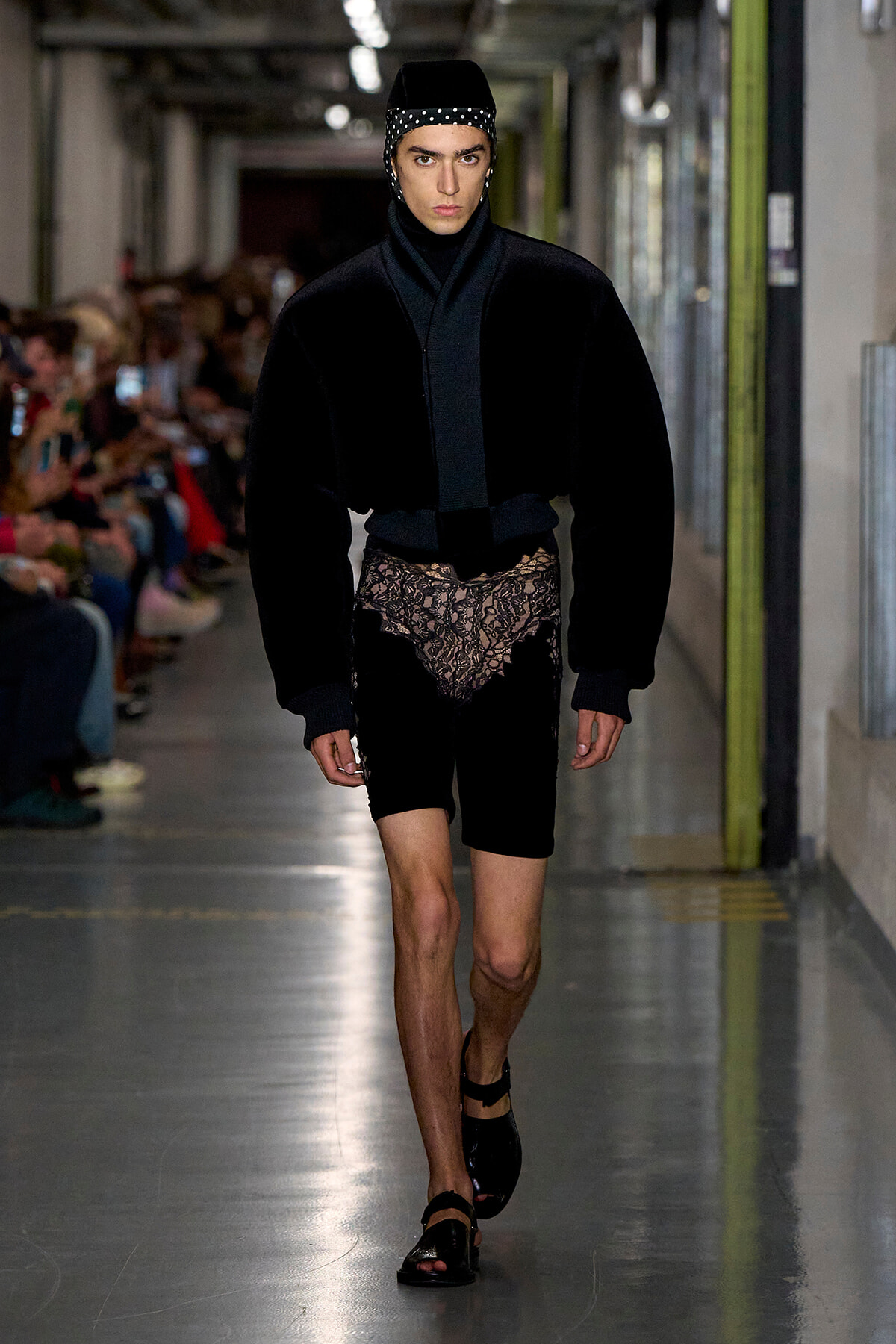 Male model walking a runway in a black velvet ensemble with lace shorts and a studded cap; audience blurred in the background.
