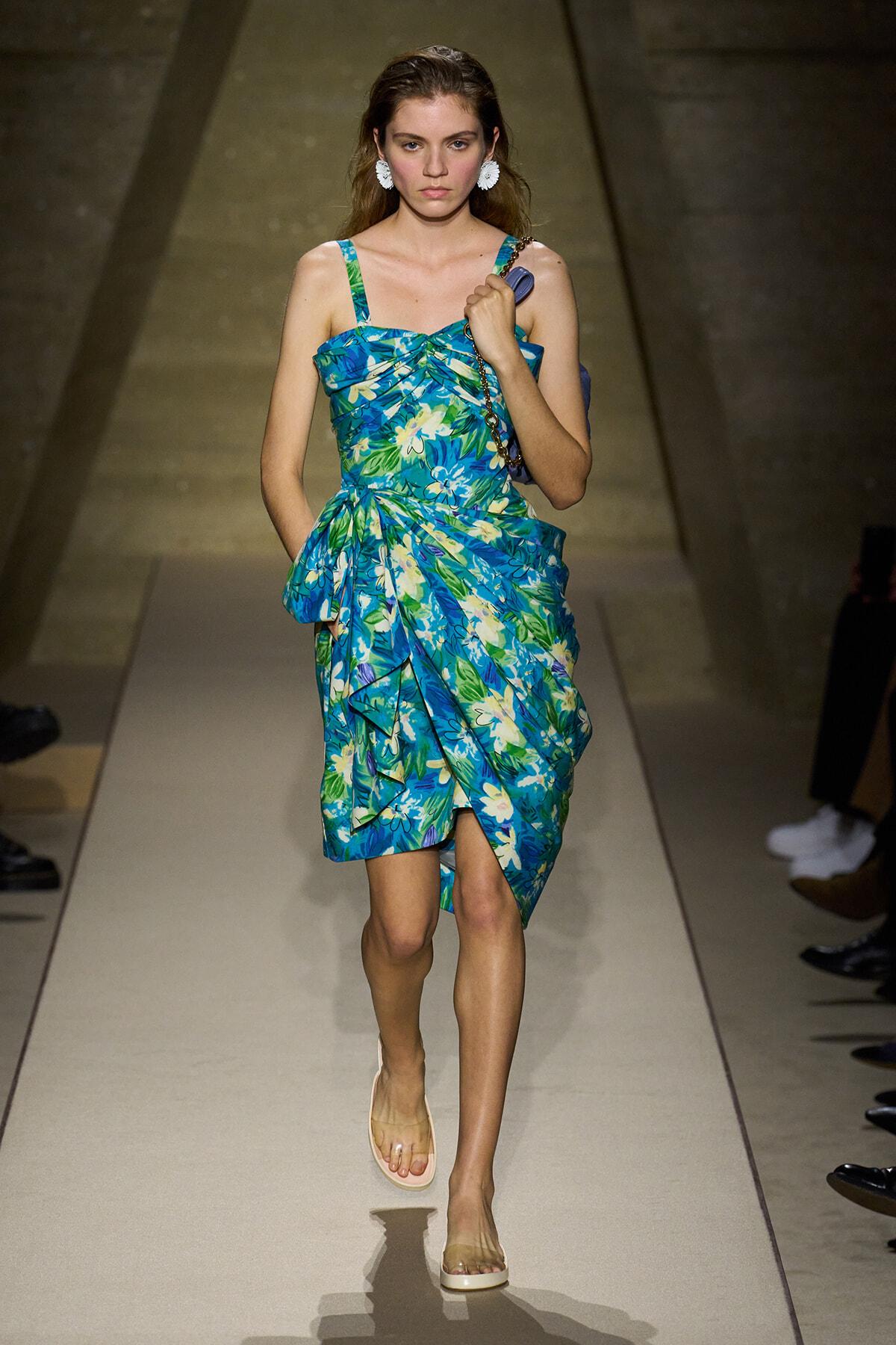 Model on a runway wearing a bright blue-green tropical floral dress with a wrapped, draped skirt and beige sandals. She has white statement earrings and carries a small bag over her shoulder.