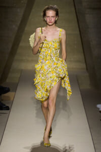 Model on a runway wearing a yellow floral print dress with a ruffled, high-low hem and matching heels, carrying a chain-strap bag.