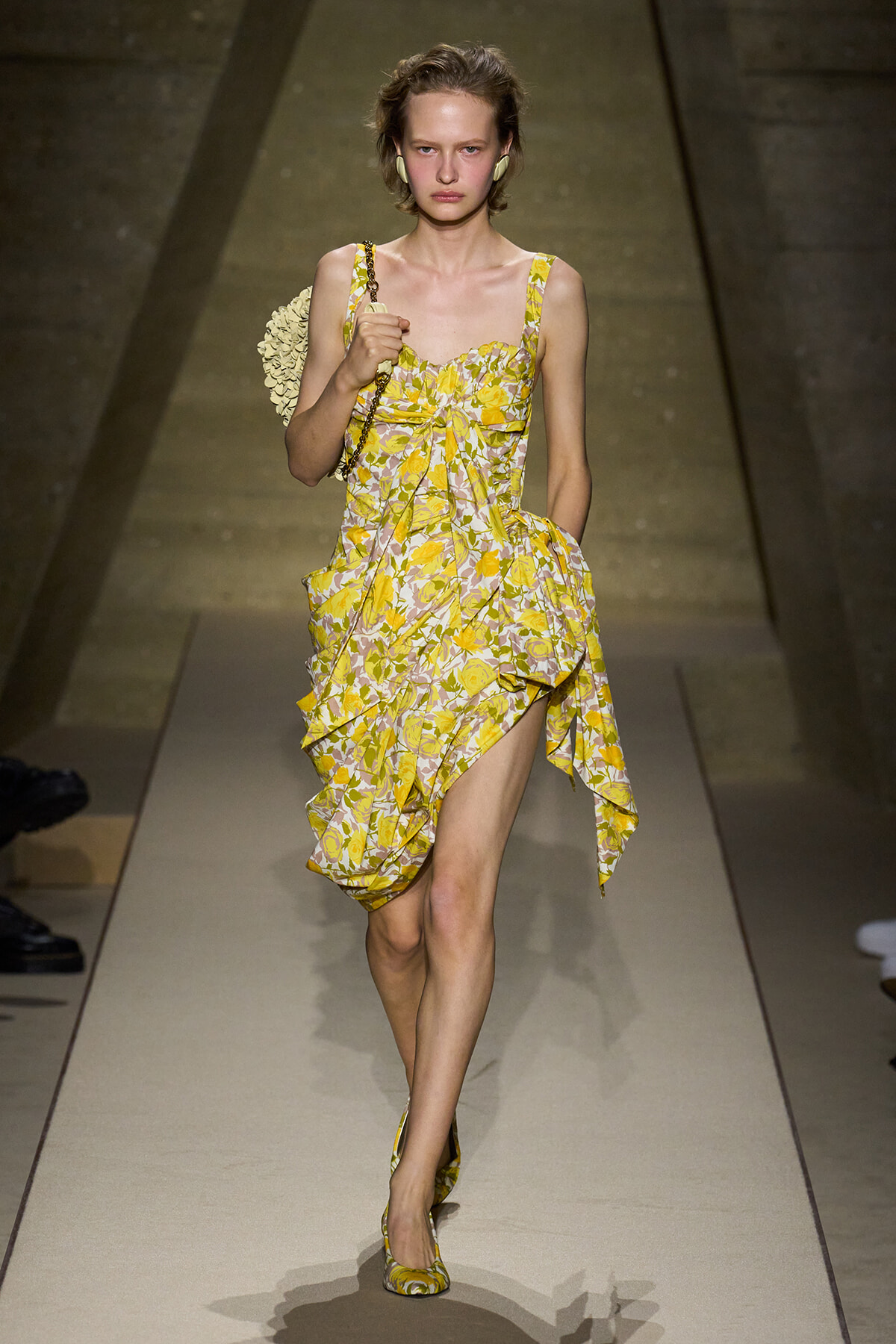 Model on a runway wearing a yellow floral print dress with a ruffled, high-low hem and matching heels, carrying a chain-strap bag.