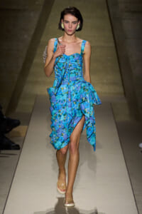 Female model walks the runway in a turquoise-blue floral-print dress with a tied front and layered, ruffled skirt, wearing clear sandals and statement earrings.