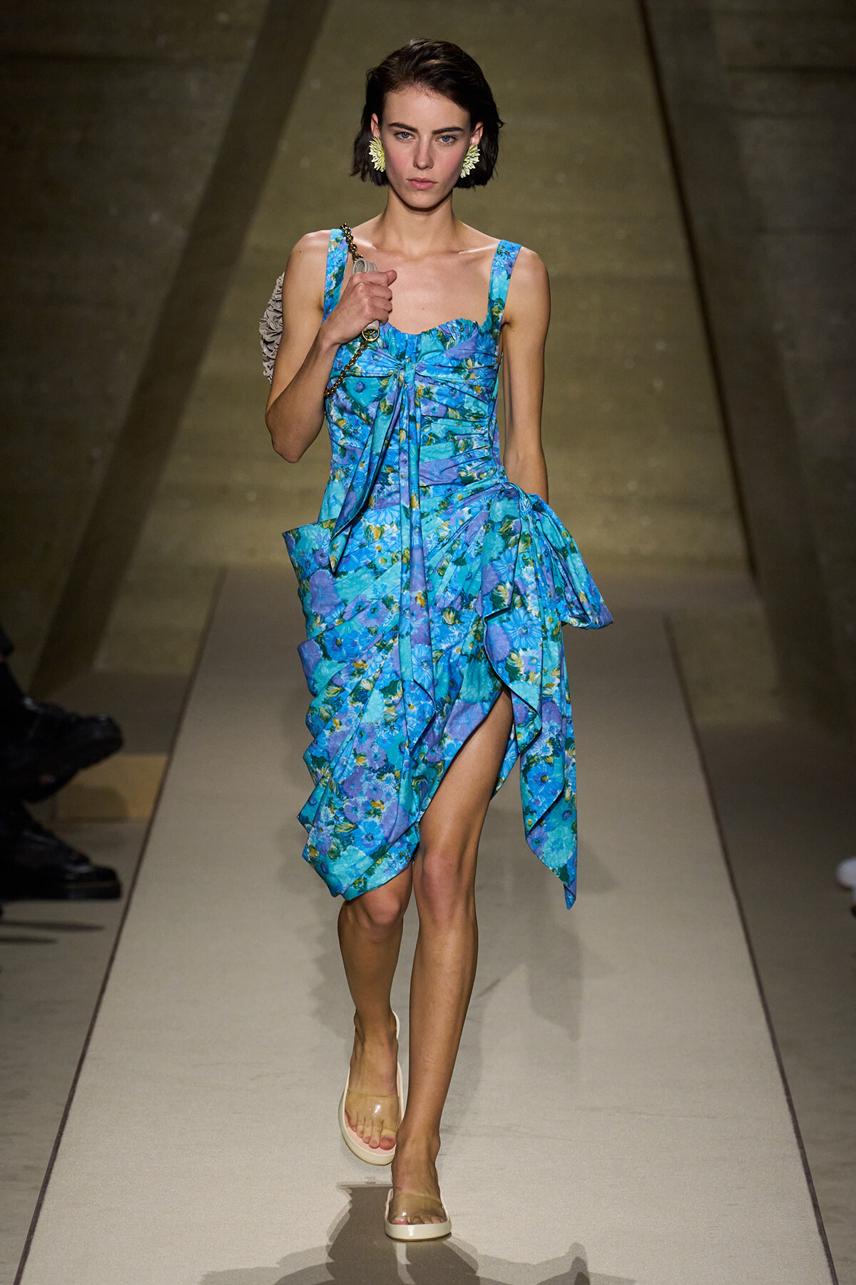 Female model walks the runway in a turquoise-blue floral-print dress with a tied front and layered, ruffled skirt, wearing clear sandals and statement earrings.