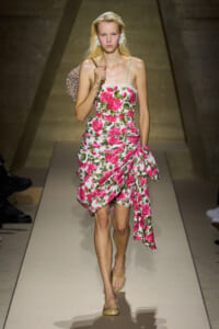 Model walks a runway in a white rose-print dress with a large pink bow and a single thin strap, carrying a chain-strap bag on her shoulder.