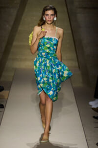 Model walking a fashion runway in a blue tropical floral dress with a yellow shoulder bag and white floral earrings.