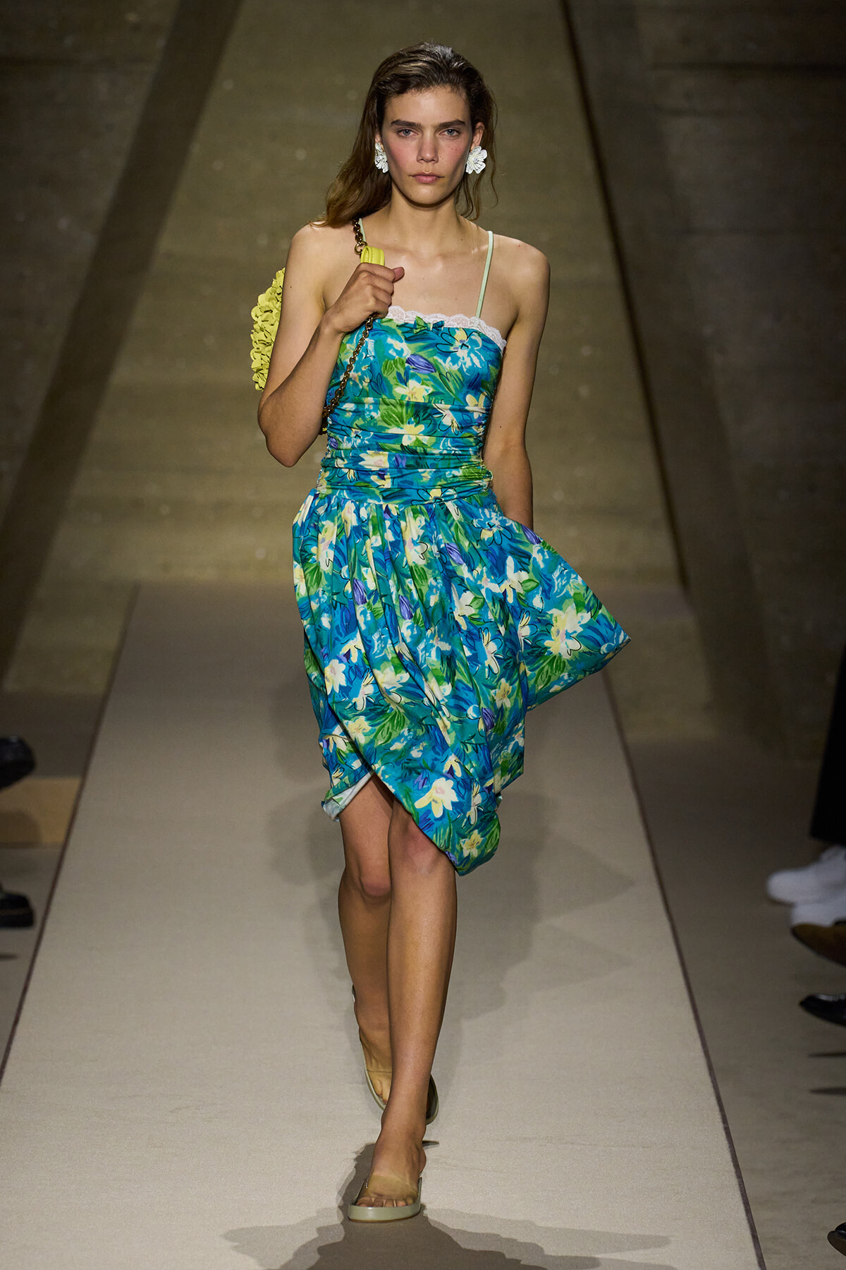 Model walking a fashion runway in a blue tropical floral dress with a yellow shoulder bag and white floral earrings.