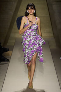 Model walking a floral purple-and-white dress on a runway with a dark back accessory and yellow heels, holding a microphone-style prop.