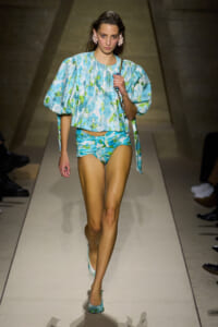 Model walking the runway in a turquoise-blue floral two-piece swimsuit with a voluminous matching top and turquoise heels, accessorized with pink floral earrings and a chain-strap bag.