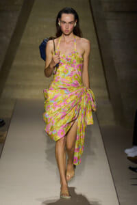 Model walks down the runway in a bright yellow floral halter dress with a draped, knotted skirt and a dark blue handbag on her shoulder.