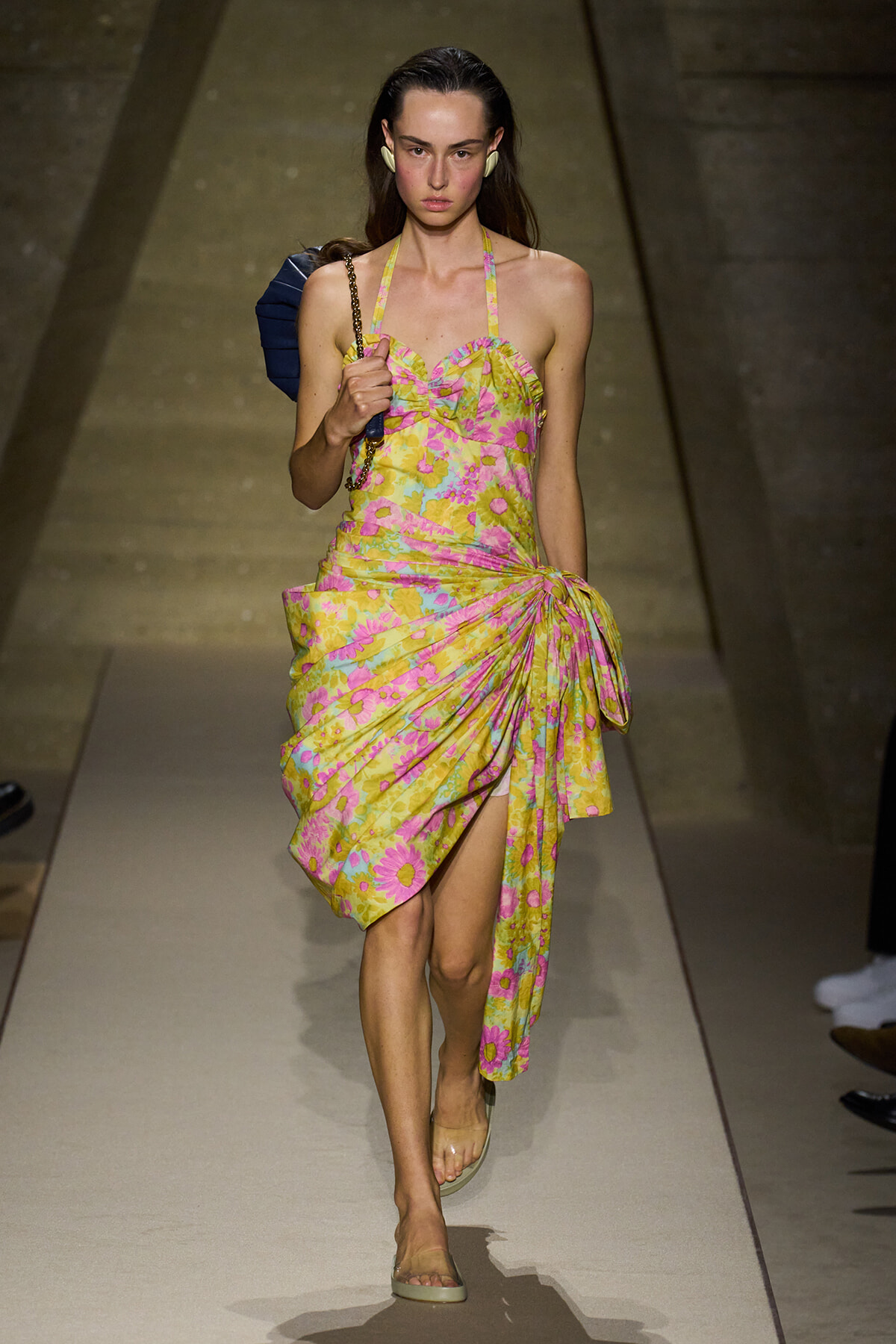 Model walks down the runway in a bright yellow floral halter dress with a draped, knotted skirt and a dark blue handbag on her shoulder.