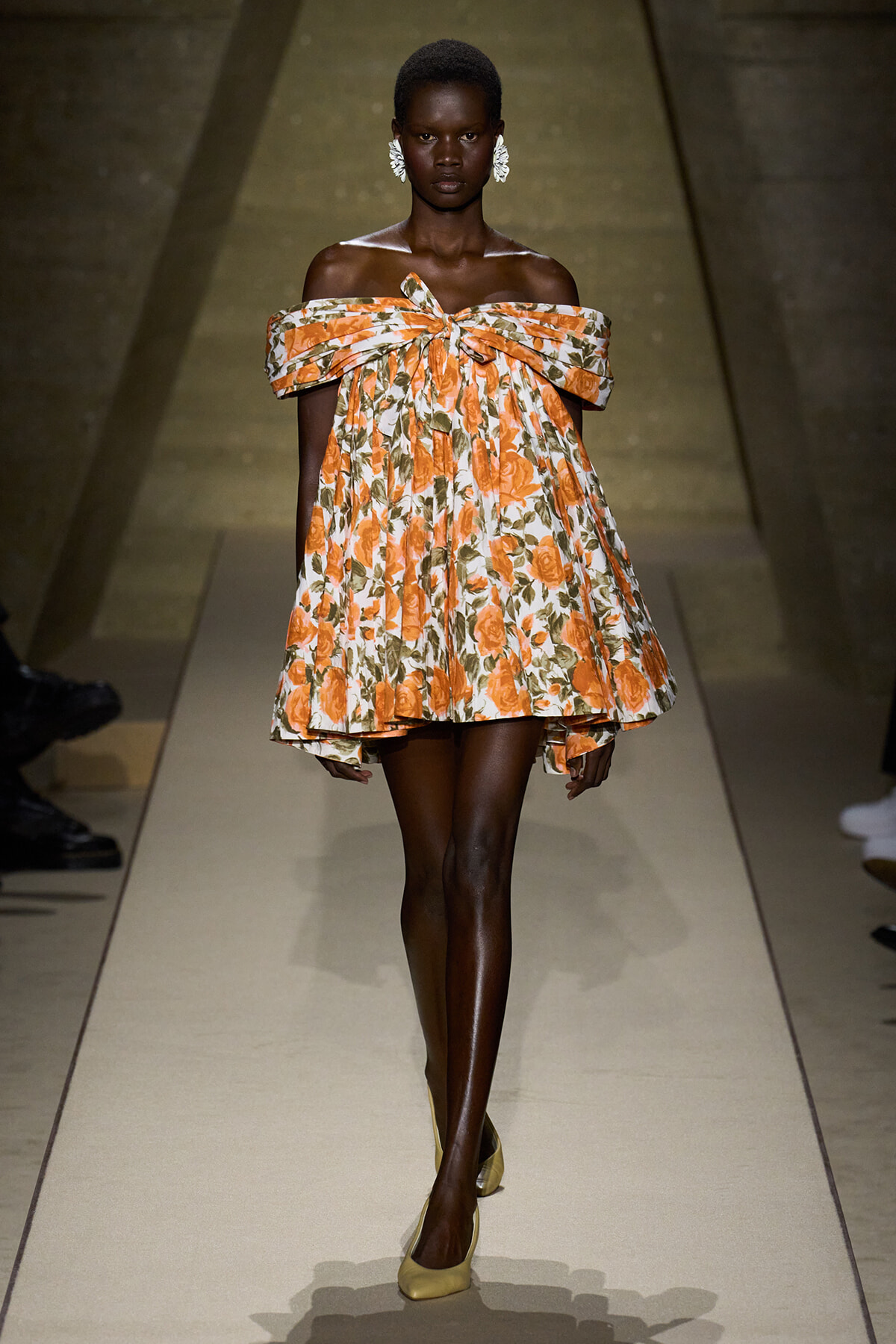 Model walking the runway in an off-the-shoulder orange floral dress with a large bow at the chest and white statement earrings.