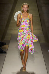 Model walking the runway in a pink-yellow floral dress with a ruched bodice and asymmetrical skirt, carrying a white circular handbag.