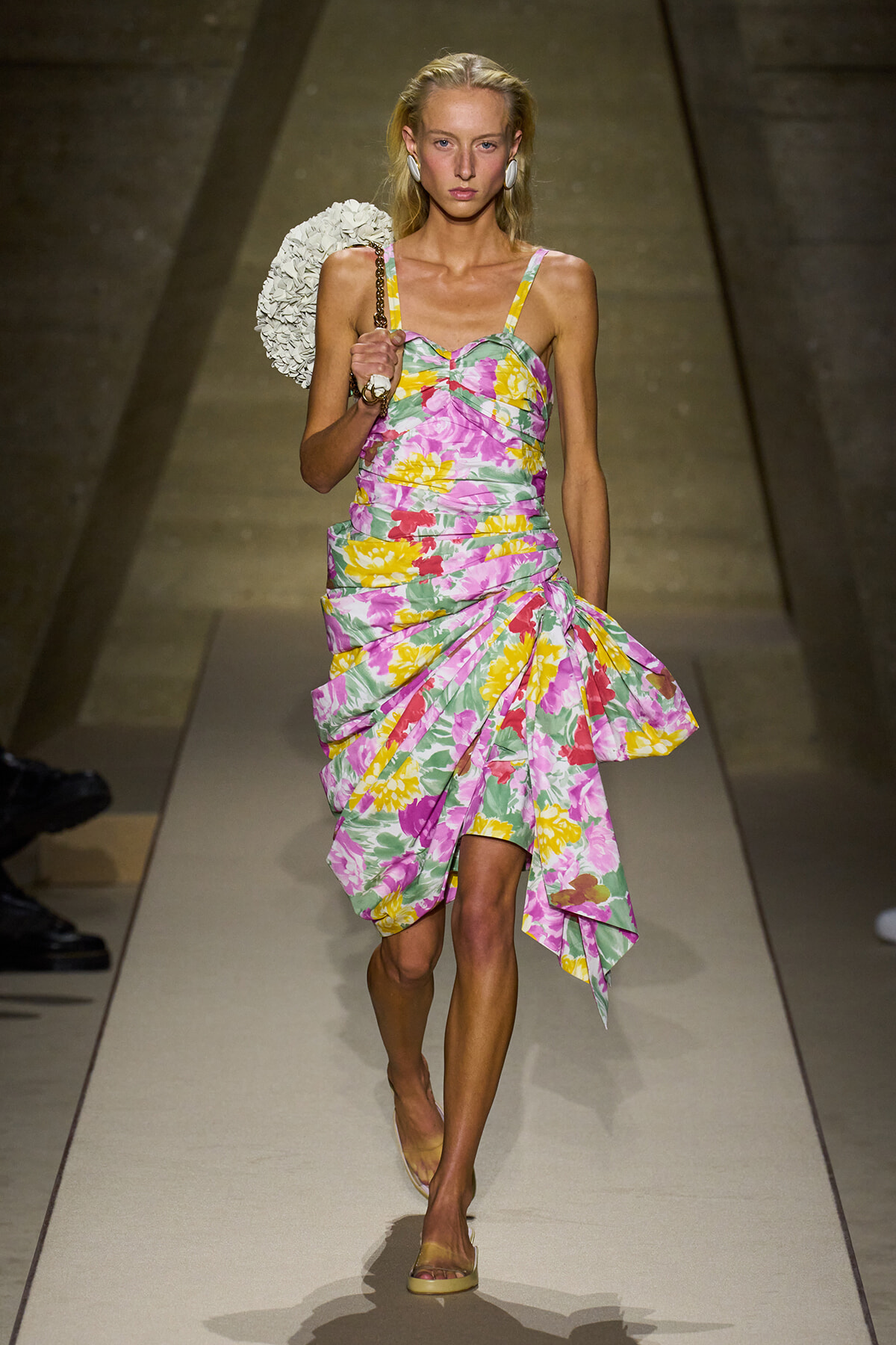 Model walking the runway in a pink-yellow floral dress with a ruched bodice and asymmetrical skirt, carrying a white circular handbag.