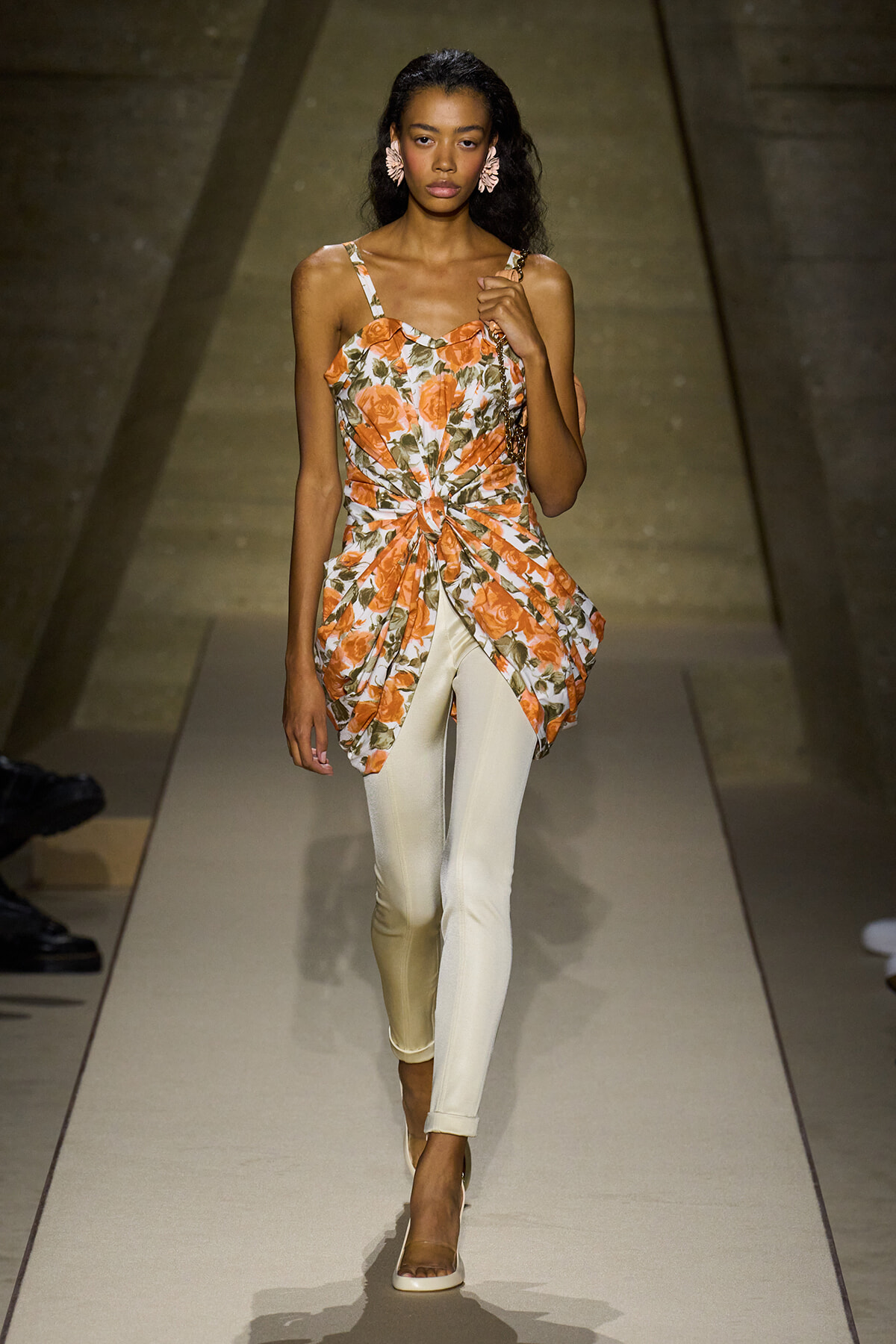 Model walks down a runway wearing an orange floral sleeveless top with a knotted front and white fitted pants, accessorized with pink flower earrings and beige heels.