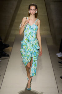 Model walking down a runway in a light blue-green floral dress with thin straps and a twisted knot at the waist, carrying a white handbag and wearing matching flats.