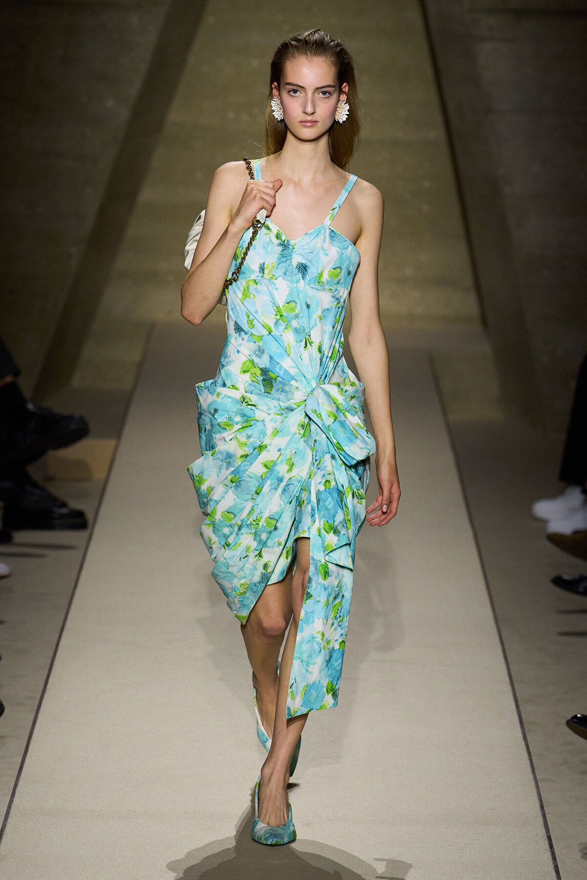 Model walking down a runway in a light blue-green floral dress with thin straps and a twisted knot at the waist, carrying a white handbag and wearing matching flats.