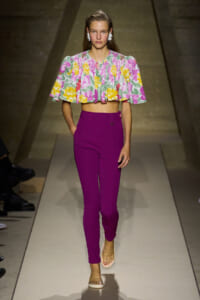 Female model walks a runway wearing a colorful floral crop top and magenta high-waisted pants. She wears beige sandals and statement earrings, walking confidently down the catwalk.
