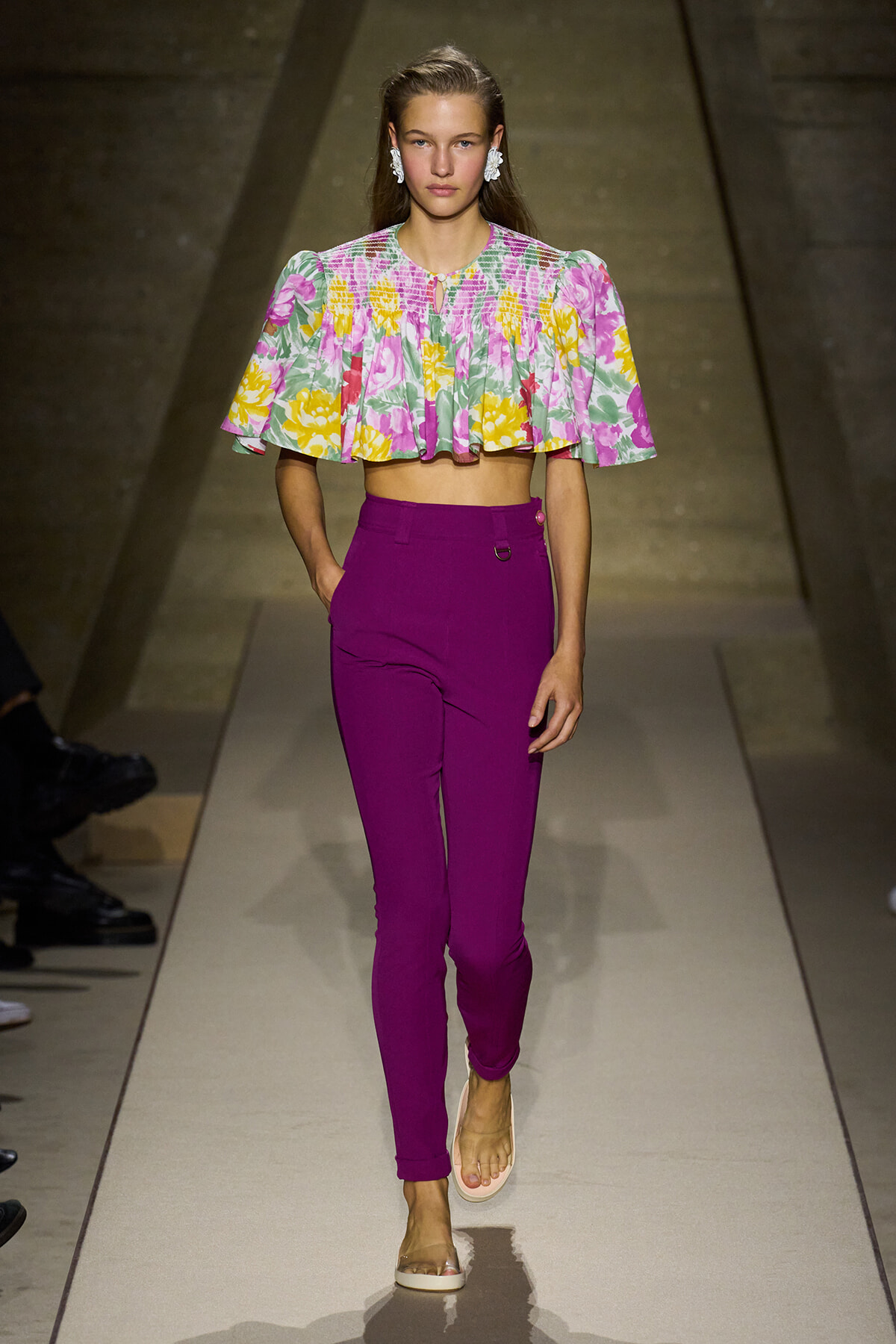 Female model walks a runway wearing a colorful floral crop top and magenta high-waisted pants. She wears beige sandals and statement earrings, walking confidently down the catwalk.