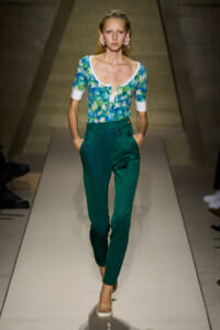 Woman model walking the runway in teal high-waisted pants and a blue-green floral top with white cuffs and neckline, accessorized with pink earrings.