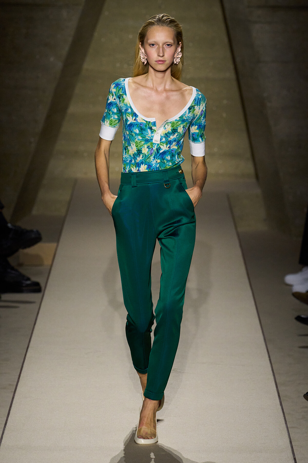 Woman model walking the runway in teal high-waisted pants and a blue-green floral top with white cuffs and neckline, accessorized with pink earrings.