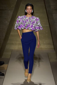 Model walking the runway in a purple floral off-the-shoulder crop top with ruffled cape sleeves and high-waisted royal blue pants, beige sandals.