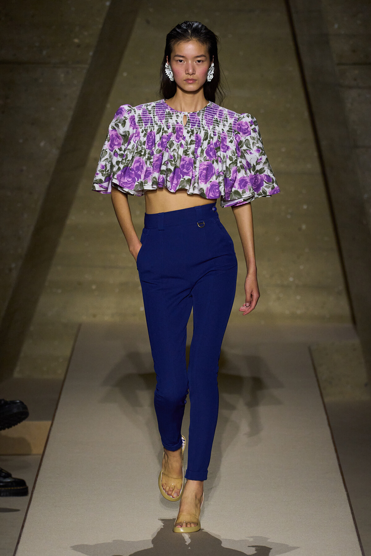 Model walking the runway in a purple floral off-the-shoulder crop top with ruffled cape sleeves and high-waisted royal blue pants, beige sandals.