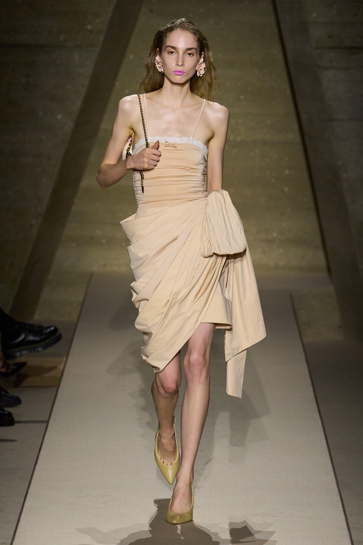 Model walking the runway in a beige draped dress with a vertical ruched bodice, lace trim at the top, and a large draped skirt, paired with yellow heels and a chain-strap handbag.