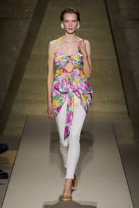 Model on a runway wearing a colorful tropical-print top with cutouts, paired with white fitted pants and yellow heels.