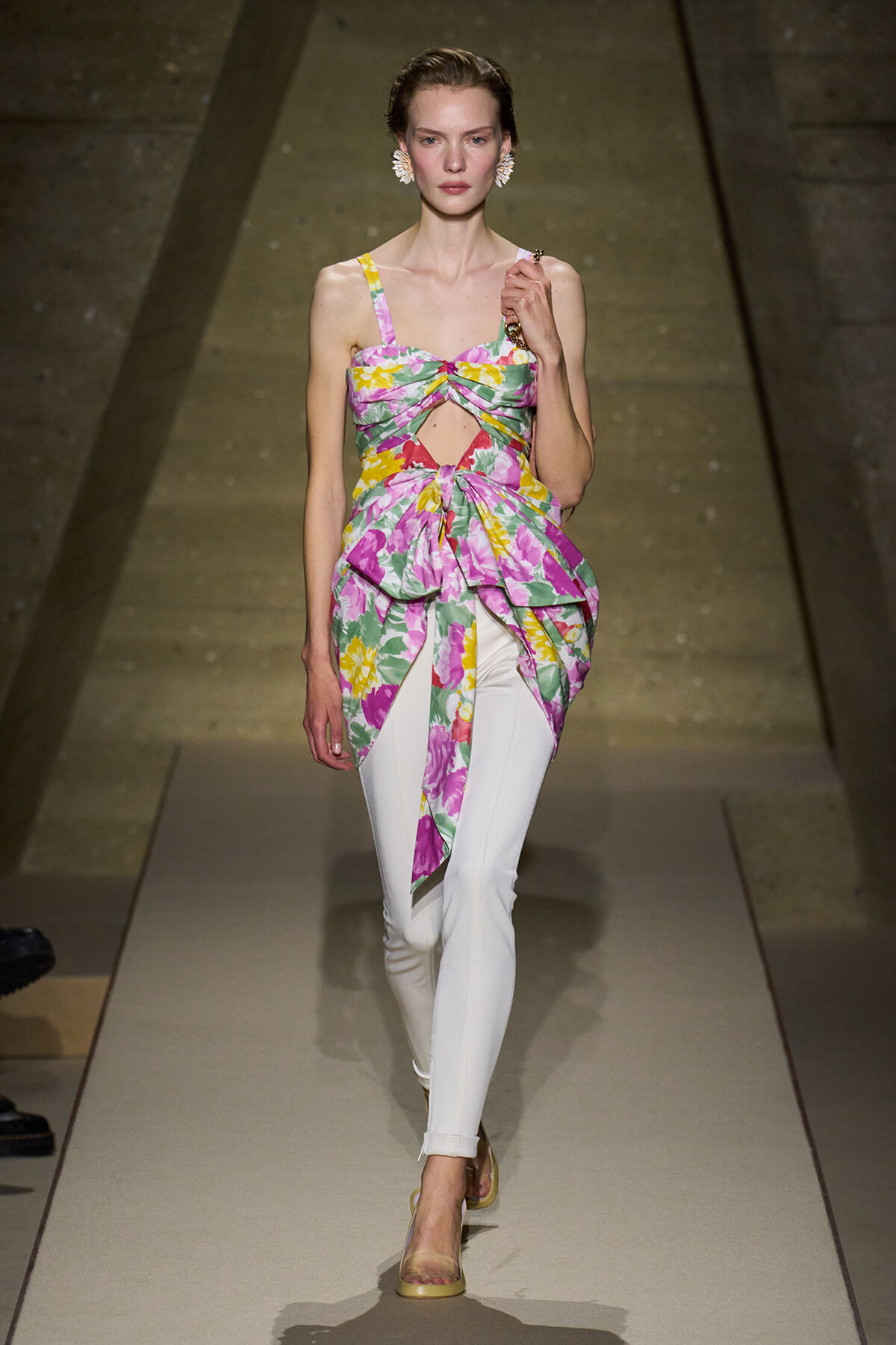 Model on a runway wearing a colorful tropical-print top with cutouts, paired with white fitted pants and yellow heels.