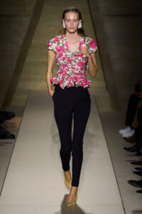 Model walking a runway in a pink floral ruched peplum top and black slim pants, beige heels, and oversized white flower earrings.