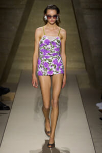 Model walking a runway in a purple floral mini dress with lace neckline and lime straps, wearing sunglasses.