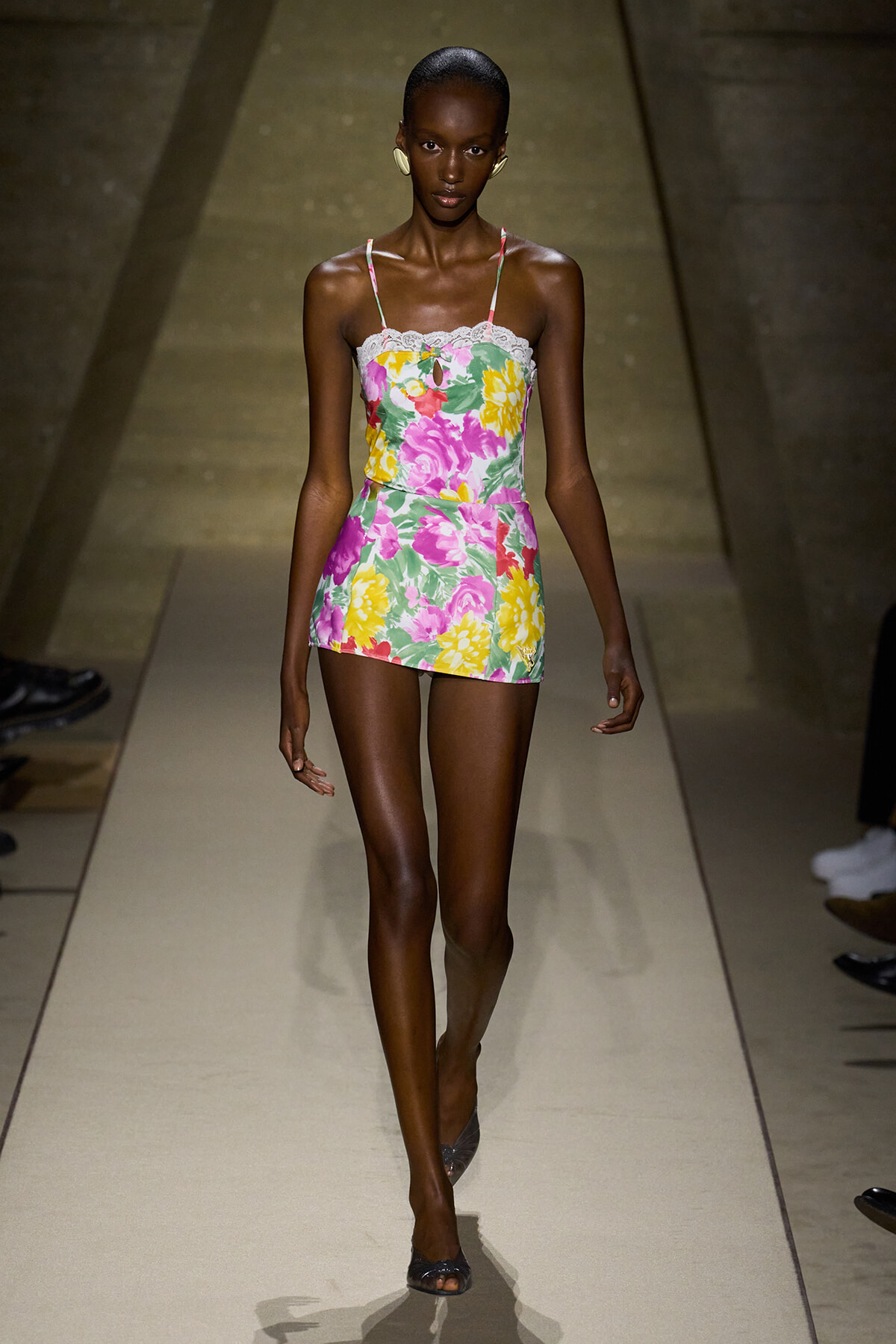 Model walking the runway in a sleeveless floral mini romper with lace-trim neckline and thin straps, bright pinks, yellows, and greens.