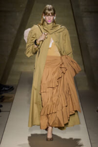 Model walking a runway in a tan trench over a layered camel dress with ruffles and an oversized bow detail at the waist, wearing nude heels and bold pink lipstick.